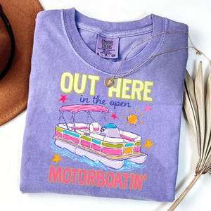 May include: A purple Comfort Colors t-shirt with a graphic of a colorful pontoon boat. The text "OUT HERE in the open MOTORBOATIN'" is printed in yellow, pink, and green. A gold necklace is draped across the shirt.