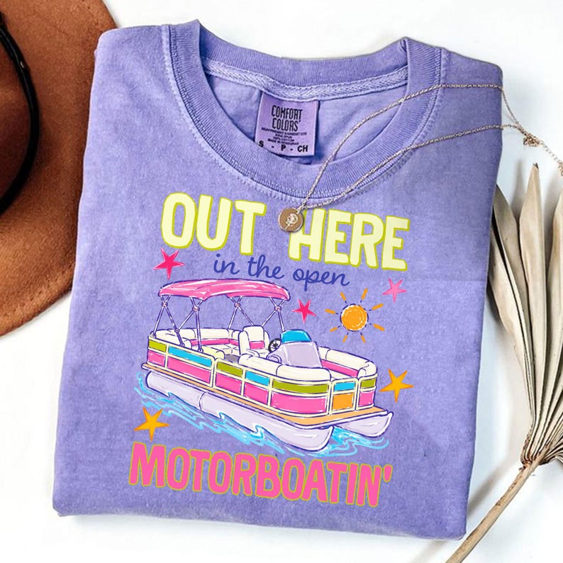 May include: A purple Comfort Colors t-shirt with a graphic of a colorful pontoon boat. The text "OUT HERE in the open MOTORBOATIN'" is printed in yellow, pink, and green. A gold necklace is draped across the shirt.