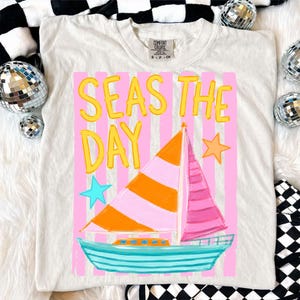 May include: Cream-colored t-shirt with a colorful graphic of a sailboat and the words "SEAS THE DAY" in yellow. The sailboat has orange and pink sails, a blue hull, and is set against pink vertical stripes. Two stars and a tag are also visible.