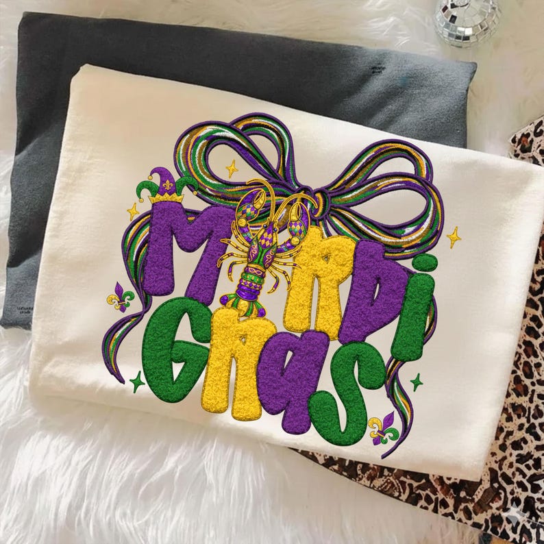 May include: Cream-colored t-shirt with a colorful Mardi Gras design. The design features the words "Mardi Gras" in green, yellow, and purple, with a bow and a crawfish. The shirt is folded on a white surface.