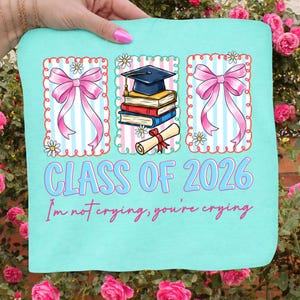 May include: A light blue graduation-themed t-shirt with the text "CLASS OF 2026" and "I'm not crying, you're crying." The design includes pink bows, daisies, a graduation cap, books, and a diploma.