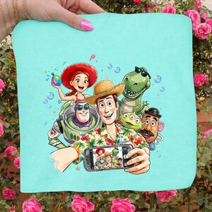 May include: A light blue square towel featuring a colorful illustration of characters from the Toy Story movie franchise taking a selfie. The characters include Woody, Buzz Lightyear, Rex, and others. The towel is held by a person.
