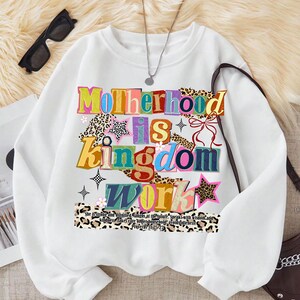 Pu&ograve; includere: Felpa bianca con la scritta "Motherhood is Kingdom Work" in lettere colorate e decorate. Il design include stelle, fiori e dettagli con stampa leopardata. Sotto &egrave; stampata una citazione da Proverbi 31:25-27.
