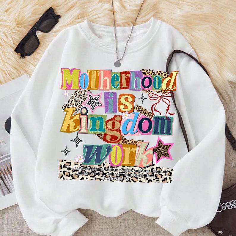 Pu&ograve; includere: Felpa bianca con la scritta "Motherhood is Kingdom Work" in lettere colorate e decorate. Il design include stelle, fiori e dettagli con stampa leopardata. Sotto &egrave; stampata una citazione da Proverbi 31:25-27.