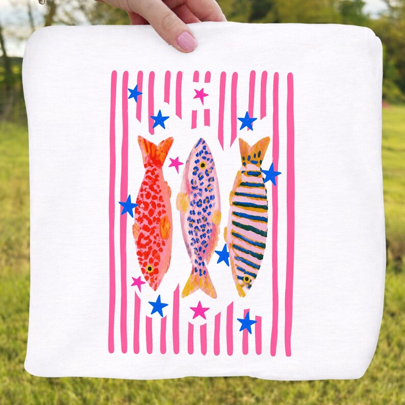 Pode incluir: Tecido quadrado branco com um design colorido de peixes. O design apresenta tr&ecirc;s peixes em padr&otilde;es vermelhos, azuis e listrados, em fundo de listras verticais rosa e estrelas azuis. O tecido &eacute; segurado por uma m&atilde;o.