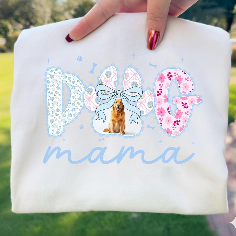 May include: White sweatshirt with the words "DOG mama" in light blue script. The letters "D" and "G" are decorated with floral patterns. The "O" in "DOG" is a paw print with a golden retriever and a blue bow.