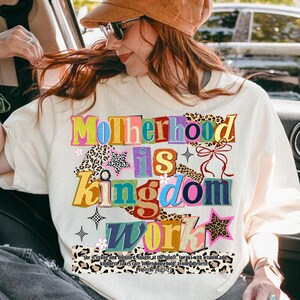 Pu&ograve; includere: T-shirt color crema con le parole "Motherhood is Kingdom Work" in lettere colorate e stratificate. Il design include stelle, fiori e dettagli con stampa leopardata. Il testo "She is strong and dignified..." &egrave; stampato sotto.