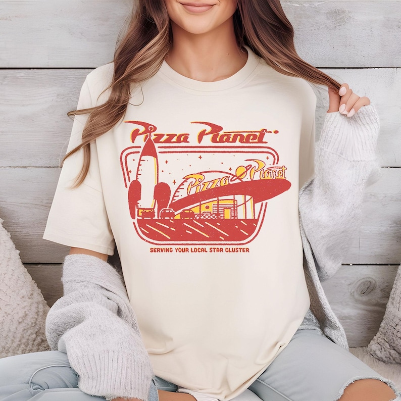 Puede incluir: Camiseta color crema con un gr&aacute;fico retro de Pizza Planet en rojo, amarillo y blanco. El dise&ntilde;o incluye un cohete, un restaurante y el texto "Pizza Planet" y "Serving Your Local Star Cluster."