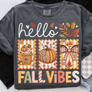 Op de afbeelding: Donkergrijs T-shirt met een herfstthema. Het ontwerp bevat het woord "hello" in witte letters boven drie rechthoekige panelen. De panelen tonen een pumpkin spice latte, een pompoen met madeliefjes en een geruite strik. De woorden "FALL VIBES" staan eronder.