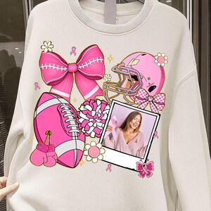 May include: Cream-colored sweatshirt featuring pink illustrations of a football, football helmet, bow, boxing gloves, and a photo of a woman holding a pink ribbon. The design includes flowers and ribbons, all in support of breast cancer awareness.