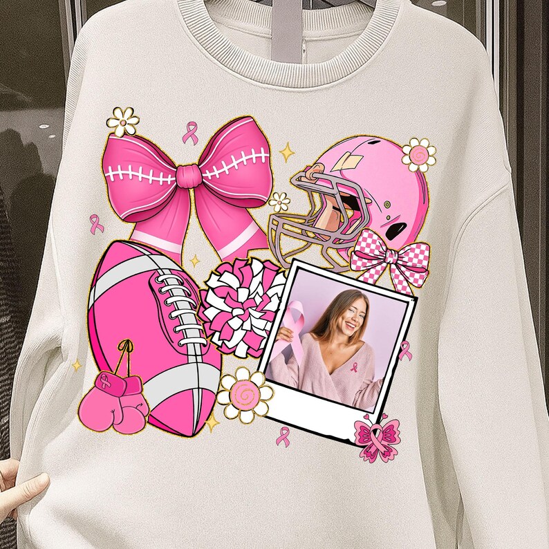May include: Cream-colored sweatshirt featuring pink illustrations of a football, football helmet, bow, boxing gloves, and a photo of a woman holding a pink ribbon. The design includes flowers and ribbons, all in support of breast cancer awareness.