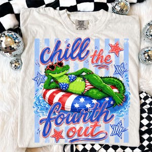 May include: Cream-colored t-shirt featuring a graphic of a green alligator wearing heart-shaped sunglasses and a patriotic bikini, lounging on a red, white, and blue inner tube. The text "chill the fourth out" is written in blue and red script.