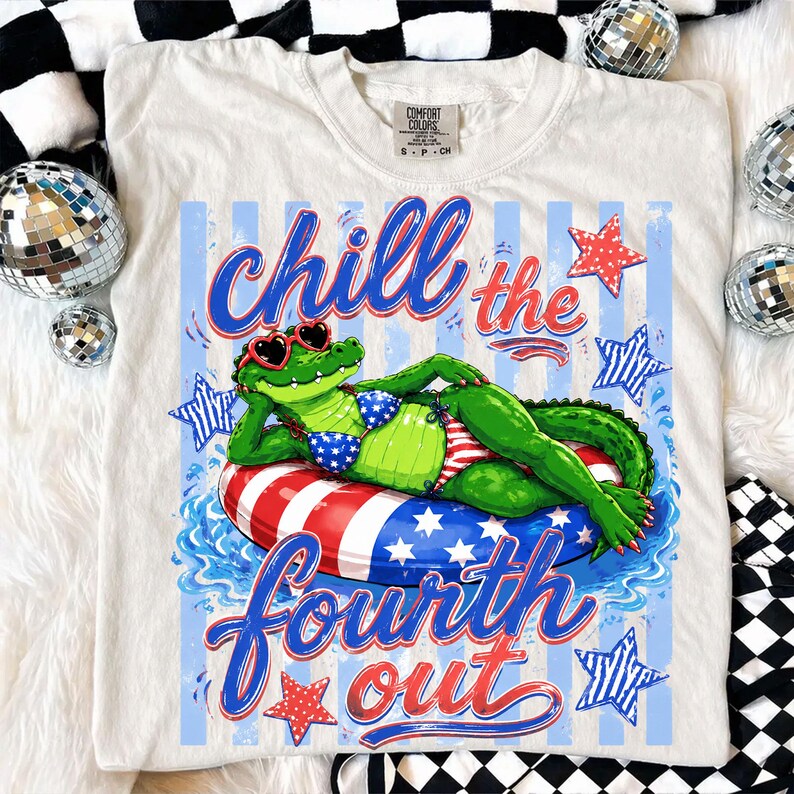 May include: Cream-colored t-shirt featuring a graphic of a green alligator wearing heart-shaped sunglasses and a patriotic bikini, lounging on a red, white, and blue inner tube. The text "chill the fourth out" is written in blue and red script.