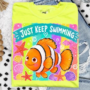 K&ouml;nnte beinhalten: Ein neongelbes T-Shirt mit einem bunten Clownfisch-Motiv und dem Text "Just Keep Swimming". Das Design umfasst Seesterne, Muscheln und einen Regenbogenhintergrund. Das Shirt ist aus einem weichen Material.