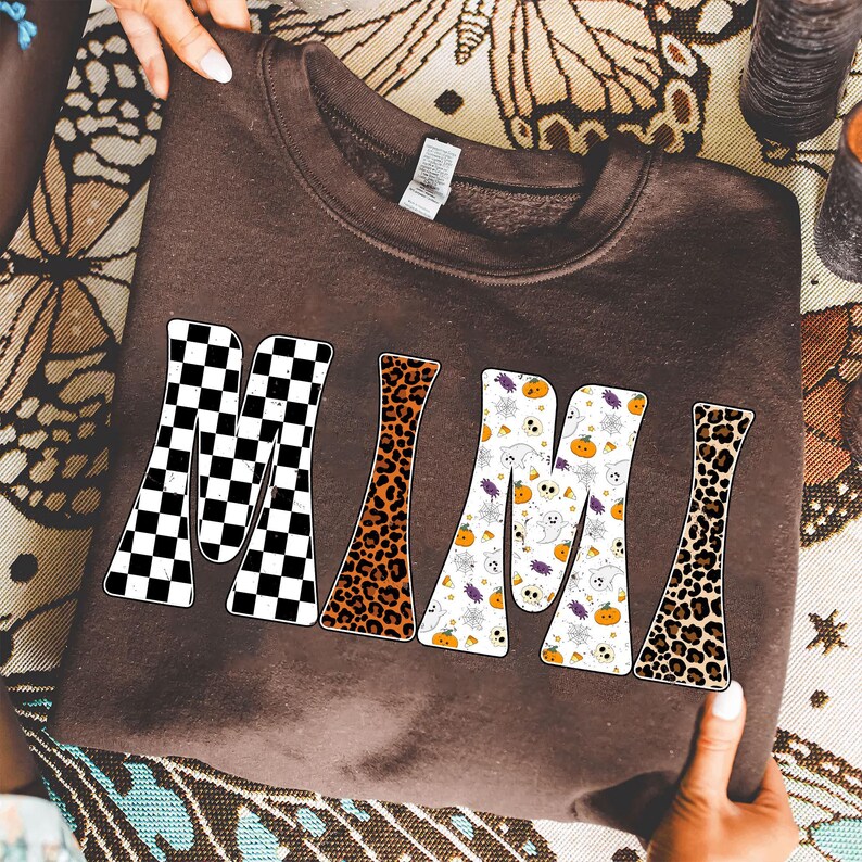 May include: A brown sweatshirt with the word "MIMI" in a retro font. The letters are patterned with black and white checkerboard, leopard print, and a Halloween-themed design with ghosts and pumpkins.