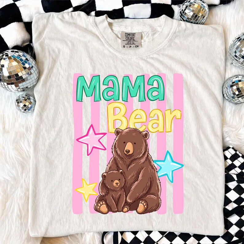 Pu&ograve; includere: T-shirt color crema con grafica "Mama Bear" in verde acqua e giallo, con un'illustrazione di un orso bruno e il suo cucciolo. Strisce rosa verticali e stelle completano il design. La maglietta &egrave; del marchio Comfort Colors.