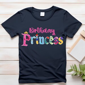 May include: A navy blue t-shirt with the text "Birthday Princess" in pink and colorful letters. The letters are decorated with images of Disney princesses.