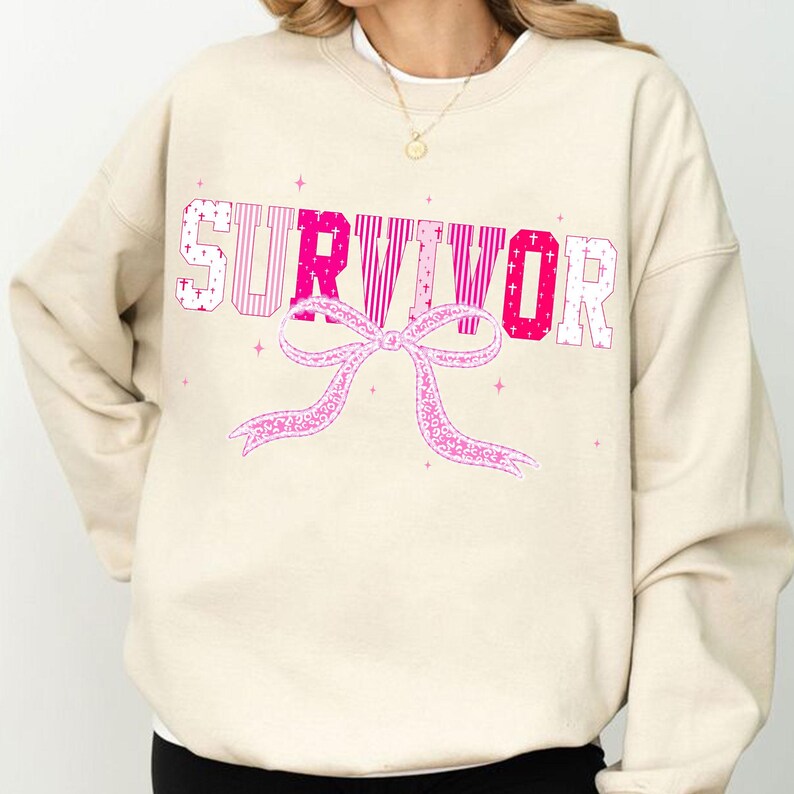 May include: A cream-colored sweatshirt with the word "SURVIVOR" in pink and white block letters. A pink bow graphic is below the word. The sweatshirt has a crew neck and long sleeves.