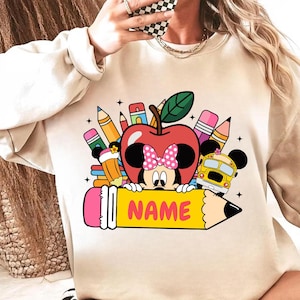 May include: Beige sweatshirt with a colorful graphic featuring Minnie Mouse as an apple, surrounded by pencils, a school bus, and the word "NAME" on a pencil. The design has a back-to-school theme.