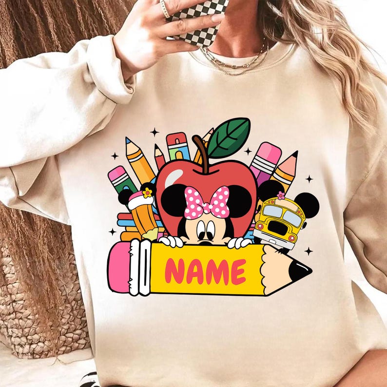 May include: Beige sweatshirt with a colorful graphic featuring Minnie Mouse as an apple, surrounded by pencils, a school bus, and the word "NAME" on a pencil. The design has a back-to-school theme.