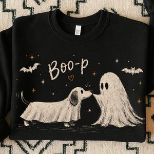 Peut inclure: Un sweat-shirt noir à col rond avec une impression graphique représentant un teckel déguisé en fantôme face à un fantôme blanc. Le texte "Boo-p" est imprimé au-dessus, accompagné de petites illustrations de chauves-souris, d'étoiles et d'un cœur.