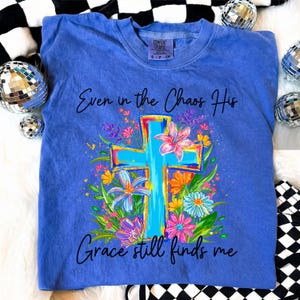 May include: A blue Comfort Colors t-shirt featuring a floral cross design with the text "Even in the Chaos His Grace still finds me." The cross is light blue with colorful accents, surrounded by flowers.