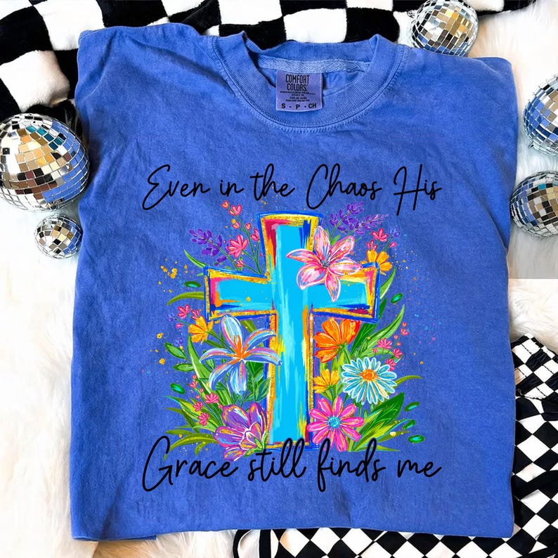 May include: A blue Comfort Colors t-shirt featuring a floral cross design with the text "Even in the Chaos His Grace still finds me." The cross is light blue with colorful accents, surrounded by flowers.