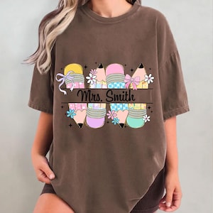 May include: Brown t-shirt featuring a colorful pencil design with bows and flowers. The text "Mrs. Smith" is displayed across the pencils. The pencils have yellow, pink, and blue accents, with black pencil tips.
