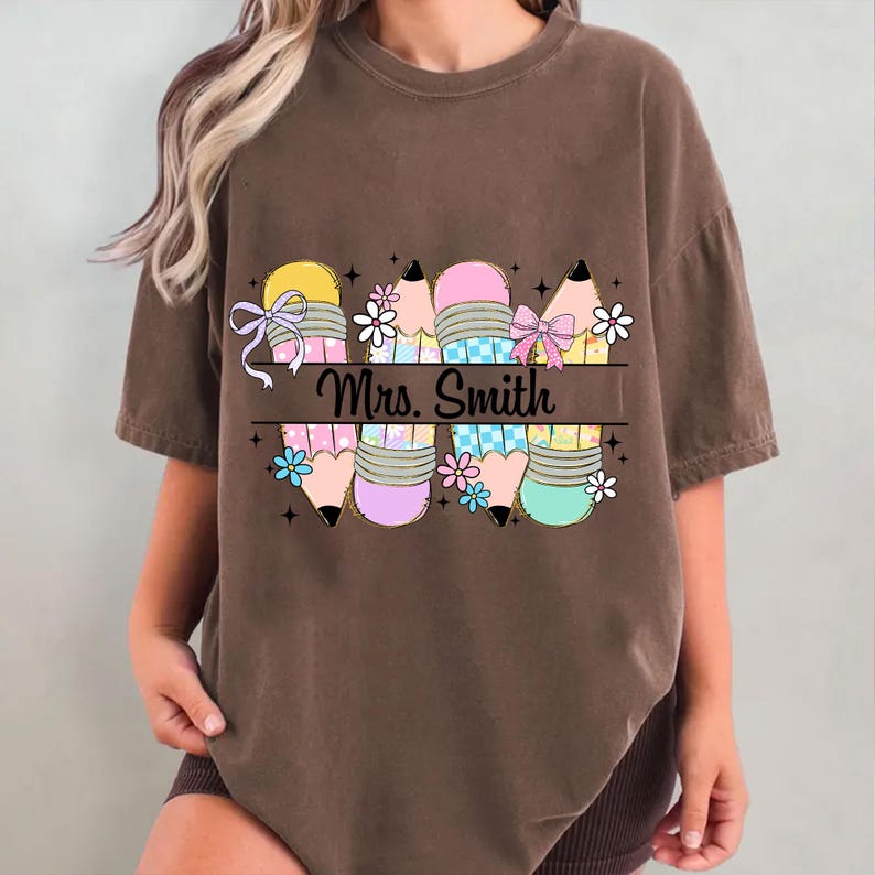 May include: Brown t-shirt featuring a colorful pencil design with bows and flowers. The text "Mrs. Smith" is displayed across the pencils. The pencils have yellow, pink, and blue accents, with black pencil tips.