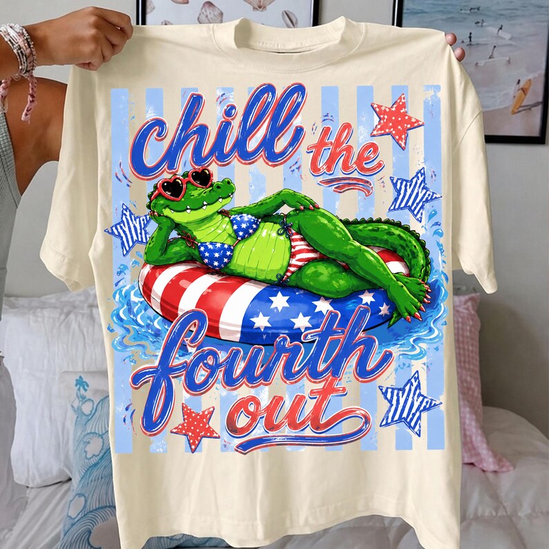 May include: Cream-colored t-shirt featuring a cartoon alligator wearing sunglasses and a bikini, lounging on a red, white, and blue inflatable ring. The shirt has the text "Chill the Fourth Out" in blue and red.
