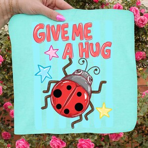 Pu&ograve; includere: Un tessuto quadrato azzurro con la scritta "GIVE ME A HUG" in rosso, insieme a una coccinella dei cartoni animati e grafiche a stella. La coccinella &egrave; rossa con pois neri e una testa grigia. Lo sfondo ha strisce verticali blu.