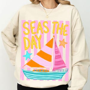 May include: Beige sweatshirt with a colorful graphic. The design features a sailboat with orange and pink sails, a blue hull, and the text "SEAS THE DAY" in yellow. Pink and white striped background with blue and orange stars.