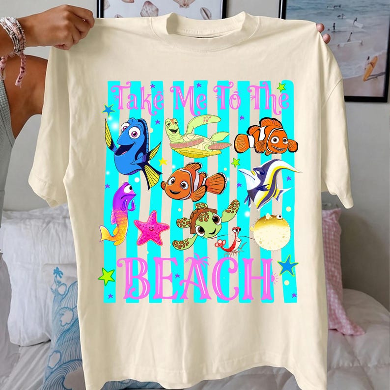 Może przedstawiać: Kremowa koszulka z kolorową grafiką przedstawiającą r&oacute;żne stworzenia morskie, w tym ryby i ż&oacute;łwie, z napisem "Take Me To The Beach". Projekt umieszczono na tle niebieskich pask&oacute;w.