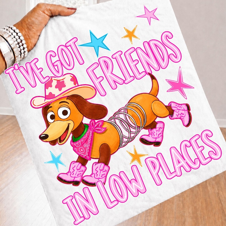 May include: A white fabric item featuring a cartoon dachshund with a metal spring body, wearing a pink cowgirl hat, a pink bandana, and pink cowboy boots. The text reads I'VE GOT FRIENDS IN LOW PLACES, surrounded by pink, blue, and orange stars.
