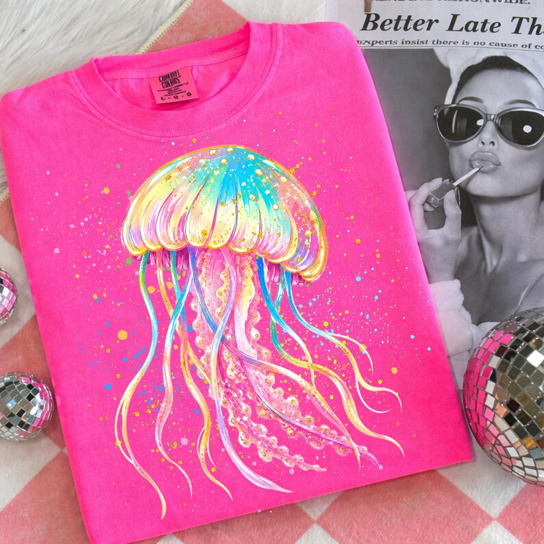 May include: A bright pink sweatshirt featuring a colorful jellyfish design. The jellyfish is depicted with a rainbow-colored bell and long, flowing tentacles. The shirt has a crew neck and long sleeves. The tag reads "Comfort Colors".