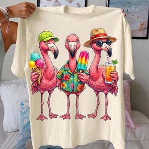 May include: Cream-colored t-shirt featuring a graphic of three pink flamingos. One flamingo wears a green cap and holds an ice cream cone. Another wears a Hawaiian shirt and holds a popsicle. The third flamingo wears sunglasses and a straw hat, holding a cocktail.