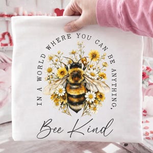 May include: White square pillow with a watercolor illustration of a bumblebee surrounded by yellow and white flowers. The text reads "In a world where you can be anything, Bee Kind."