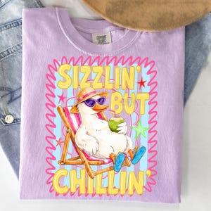 May include: A lilac Comfort Colors t-shirt featuring a cartoon duck in sunglasses and a straw hat, relaxing in a beach chair with a coconut drink. The shirt has the text "Sizzlin' But Chillin'" in yellow and pink.