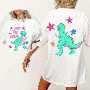 Może przedstawiać: Biała koszulka z kresk&oacute;wkowym dinozaurem w kolorze turkusowym i r&oacute;żowym, z napisem "OUT THE DOOR" i grafiką gwiazd. Tył koszulki r&oacute;wnież przedstawia dinozaura i gwiazdy. Swobodny, zabawny design.