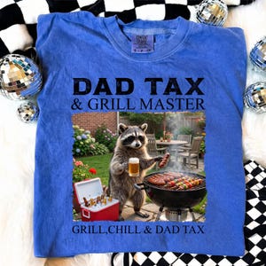 May include: Blue Comfort Colors sweatshirt with the text "DAD TAX & GRILL MASTER" and a graphic of a raccoon grilling. The raccoon is holding a beer and a sausage. The text "GRILL, CHILL & DAD TAX" is also on the shirt.