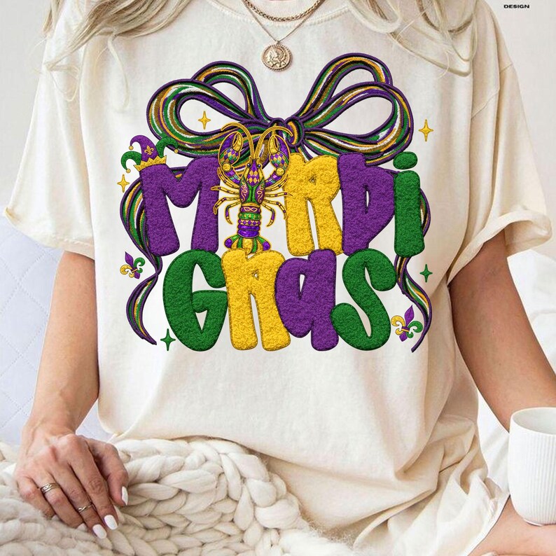 May include: Cream-colored t-shirt featuring the words "Mardi Gras" in large, colorful letters. The design includes a purple bow, a crawfish, and decorative elements in green, yellow, and purple, evoking a festive atmosphere.