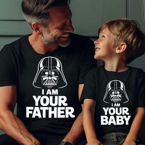 May include: Black t-shirts with white graphic of Darth Vader's helmet. The adult's shirt says "I AM YOUR FATHER," while the child's shirt says "I AM YOUR BABY." The shirts are worn by a father and child.