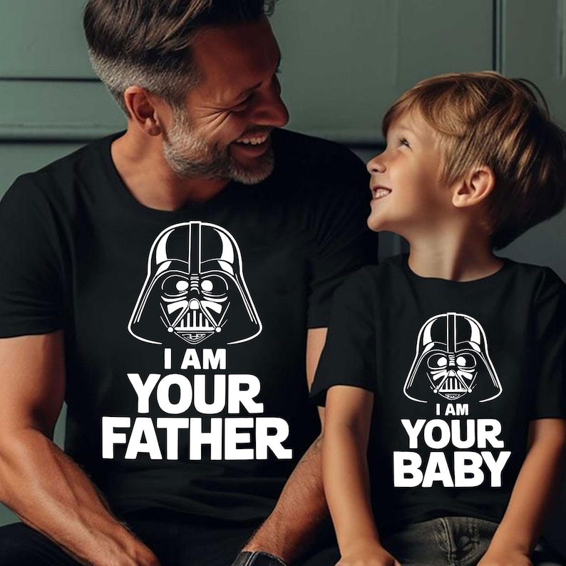 May include: Black t-shirts with white graphic of Darth Vader's helmet. The adult's shirt says "I AM YOUR FATHER," while the child's shirt says "I AM YOUR BABY." The shirts are worn by a father and child.