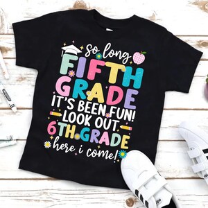Puede incluir: Camiseta negra con texto colorido: "So long FIFTH GRADE, It's been fun! Look out 6TH GRADE, here I come!" Presenta un gorro de graduaci&oacute;n, una manzana y gr&aacute;ficos de flores. Zapatillas blancas visibles en la esquina inferior derecha.