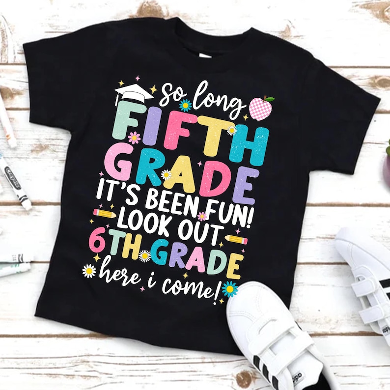 Puede incluir: Camiseta negra con texto colorido: "So long FIFTH GRADE, It's been fun! Look out 6TH GRADE, here I come!" Presenta un gorro de graduaci&oacute;n, una manzana y gr&aacute;ficos de flores. Zapatillas blancas visibles en la esquina inferior derecha.