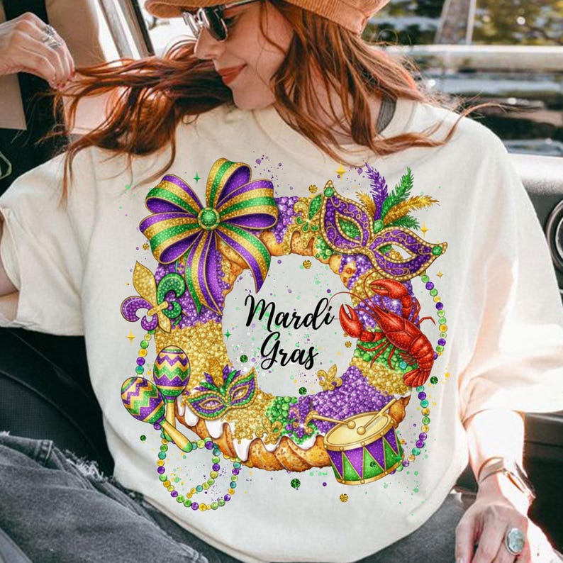 May include: Cream-colored sweatshirt featuring a vibrant Mardi Gras wreath design. The wreath includes a purple and green bow, a mask, a red crawfish, a drum, and maracas. The words "Mardi Gras" are written in black script within the wreath.