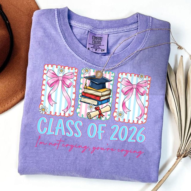 May include: A lilac Comfort Colors t-shirt with a graduation theme. The shirt features three framed designs with pink bows, a graduation cap, books, and a diploma. The text "CLASS OF 2026" and "I'm not crying, you're crying" are printed on the shirt.