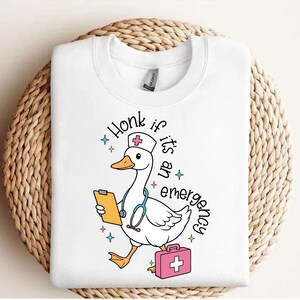 Puede incluir: Camiseta blanca con un ganso de dibujos animados que lleva un gorro de enfermera y sostiene un portapapeles. El texto "Honk if it's an emergency" est&aacute; escrito alrededor del ganso.