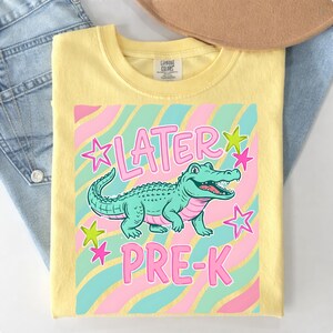 May include: Yellow t-shirt with a graphic of a teal alligator and the words "LATER PRE-K" in pink. The design includes pink and teal stripes and star accents. The shirt is folded on top of blue jeans.