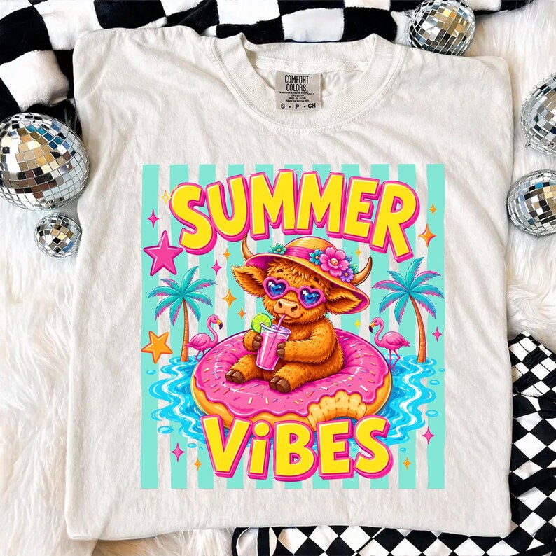 May include: Cream-colored t-shirt featuring a colorful graphic. The graphic includes the words "SUMMER VIBES" in yellow and pink, a cartoon Highland cow wearing sunglasses and a hat, and a pink donut float. Palm trees and flamingos are also included.